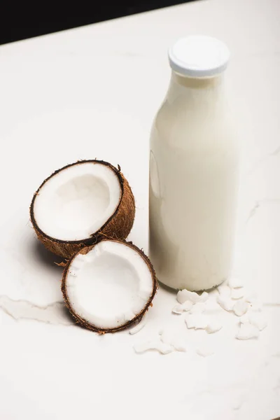 Botella de leche de coco cerca de mitades y copos en la superficie blanca aislado en negro - foto de stock