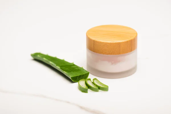 Foglia di aloe vera vicino a fette e contenitore di crema cosmetica fatta in casa su superficie bianca — Foto stock