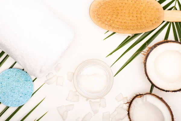 Vista superior de metades de coco e flocos, creme cosmético, toalha e escova de massagem perto de folhas de palma em branco — Fotografia de Stock