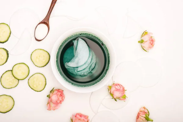 Draufsicht auf Schüssel mit Tonikum und Wattepads, Teerosen, frischen Gurkenscheiben und Löffel mit Kosmetikcreme auf Weiß — Stockfoto