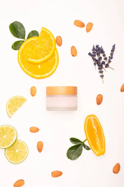 Vista superior de la crema cosmética casera cerca de naranja en rodajas y lima, almendras, ramitas de lavanda y hojas de rosa en blanco - foto de stock