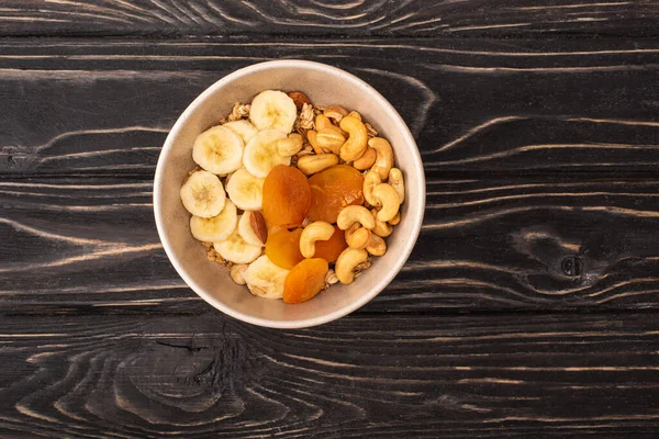 Blick von oben auf köstliches Müsli mit Nüssen, Bananen und getrockneten Aprikosen auf schwarzer Holzoberfläche — Stockfoto