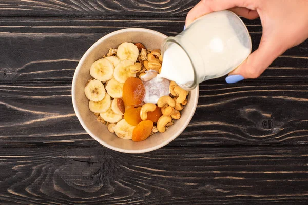 Vista cortada da mulher adicionando iogurte a granola deliciosa com nozes, banana e damascos secos na superfície preta de madeira — Fotografia de Stock