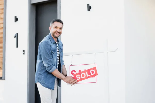 Fröhlicher Mann blickt in Kamera und hält Schild mit verkauftem Schriftzug in der Nähe des Hauses — Stockfoto