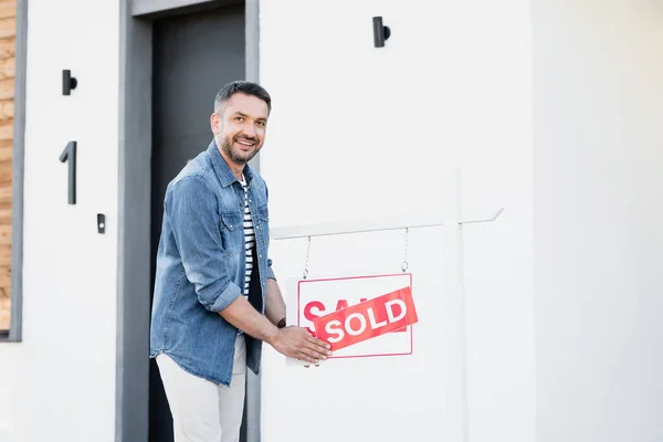 Homme joyeux regardant la caméra tout en tenant signe avec lettrage vendu près de la maison — Photo de stock