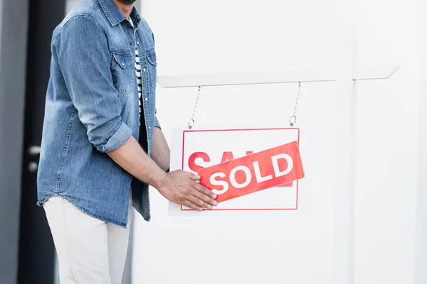 Vista ritagliata dell'uomo che tiene il segno con la scritta venduta vicino alla casa su sfondo sfocato — Foto stock