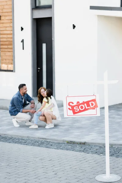 Signe avec lettrage vendu avec famille heureuse floue près de la maison sur fond — Photo de stock