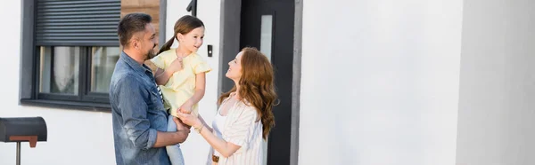 Smiling mother looking at daughter in arms of father near house, banner — Stock Photo