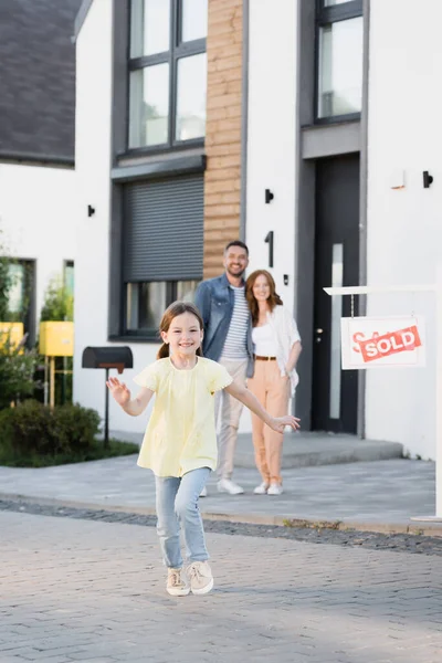 Glückliche Tochter läuft mit verschwommenem Vater und Mutter, die sich im Hintergrund in der Nähe des Hauses umarmen — Stockfoto