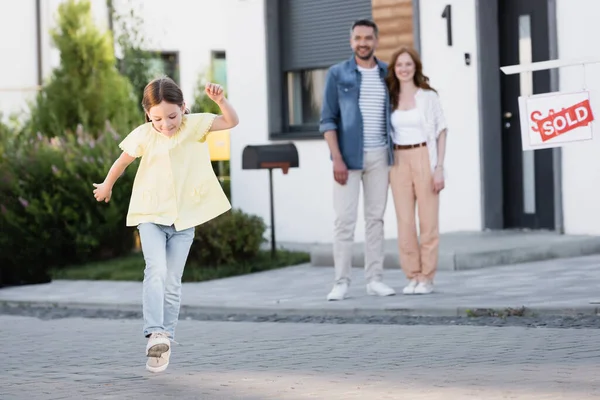 Sorrindo menina correndo com pai desfocado e mãe abraçando no fundo perto de casa — Fotografia de Stock