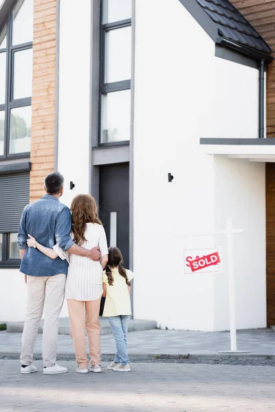 Full length of daughter standing near father and mother hugging near house and sign with sold lettering — Stock Photo