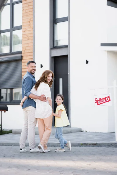 Toute la longueur de la famille souriante avec fille regardant la caméra tout en se tenant près de la maison et signe avec lettrage vendu — Photo de stock