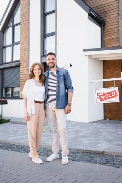 In voller Länge lächelndes Paar, das sich umarmt und in die Kamera schaut, neben Schild mit verkauftem Schriftzug und Haus — Stockfoto