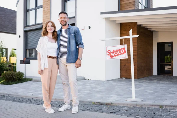 In voller Länge ein glückliches Paar, das in die Kamera schaut und sich umarmt, während es neben einem Schild mit dem Schriftzug und dem Haus steht — Stockfoto