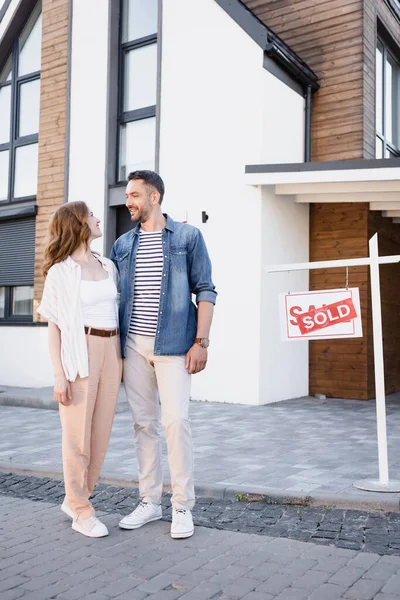 In voller Länge lächelndes Paar, das sich beim Umarmen in der Nähe von Haus und Schild mit verkauftem Schriftzug anschaut — Stockfoto