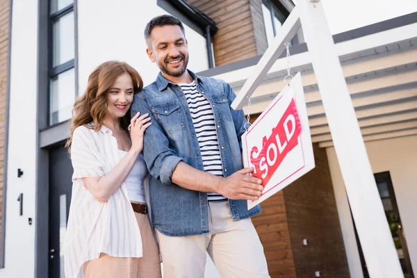 Fröhliches Paar schaut auf Schild mit verkauftem Schriftzug in der Nähe von Haus — Stockfoto