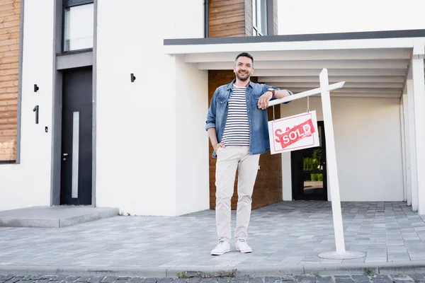 Comprimento total do homem sorridente com a mão no bolso inclinando-se no sinal com lettering vendido perto da casa — Fotografia de Stock