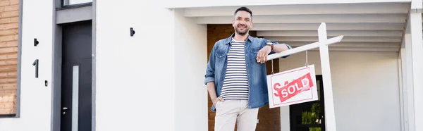 Happy man with hand in pocket looking at camera while leaning on sign with sold lettering, banner — Stock Photo