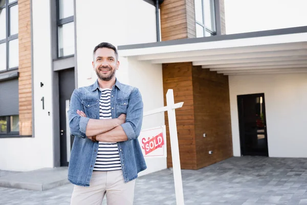 Happy man with crossed arms looking at camera near house and sign with sold lettering — Stock Photo
