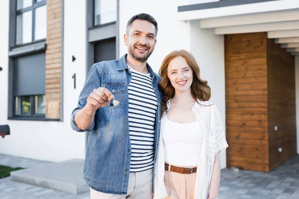 Happy man with key hugging woman while looking at camera with blurred house on background — Stock Photo