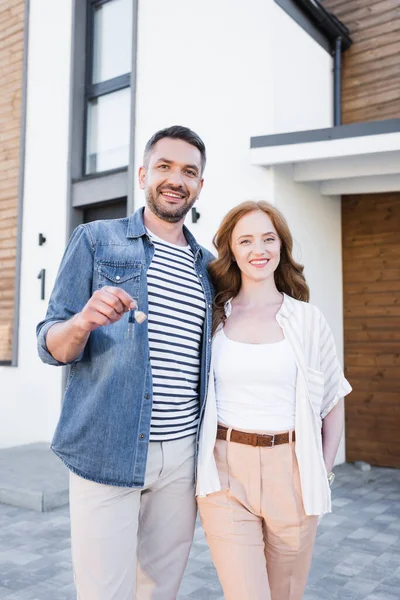 Mujer feliz abrazando hombre con llave y mirando a la cámara cerca de casa - foto de stock