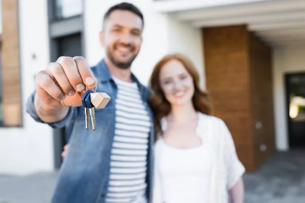 Ключи и брелок в руке счастливого мужчины обнимающего женщину возле дома на размытом фоне — стоковое фото