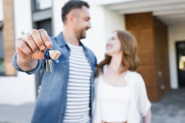 Ключи и брелок в руках счастливого мужа, смотрящего и обнимающего жену возле дома на размытом фоне — стоковое фото