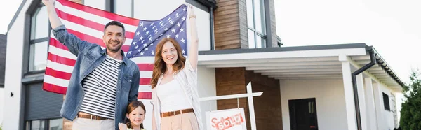Pareja feliz con hija mirando a la cámara mientras sostiene la bandera americana cerca del letrero con letras y casa vendidas, pancarta - foto de stock