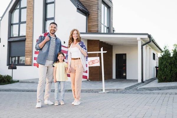 Volle Länge der glücklichen Tochter mit Mama und Papa, die eine amerikanische Flagge halten, während sie zusammen stehen und in die Kamera in der Nähe des Hauses schauen — Stockfoto