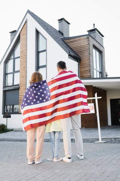 Back view of daughter with mom and dad covered with american flag while standing together near house — Stock Photo