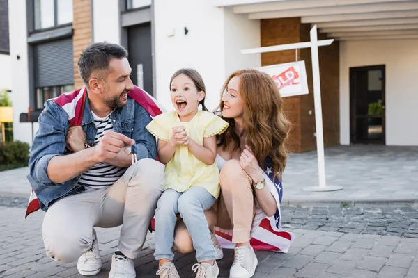 Aufgeregte Tochter mit offenem Mund sitzt neben Mutter und Vater und zeigt Schlüssel mit verschwommenem Haus im Hintergrund — Stockfoto