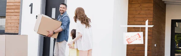 Marido sorridente com caixa de papelão olhando para a esposa de mãos dadas com a filha perto de casa e assinar com letras vendidas, banner — Fotografia de Stock