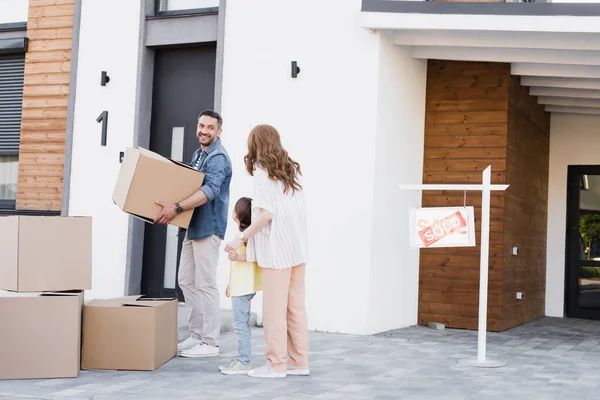 Mari souriant avec boîte en carton regardant femme tenant la main avec fille près de la maison et signe avec lettrage vendu — Photo de stock