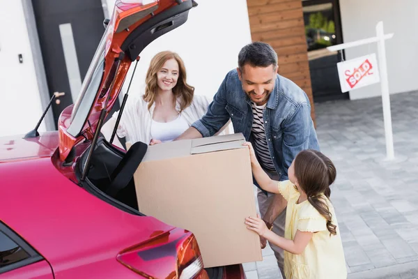 Sonriente padre mirando a la hija mientras toma la caja de cartón del maletero del coche cerca de la esposa con la casa borrosa en el fondo - foto de stock