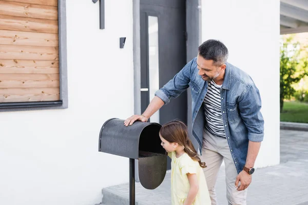 Sorridente padre in piedi vicino a figlia guardando nella cassetta della posta vuota vicino a casa — Foto stock