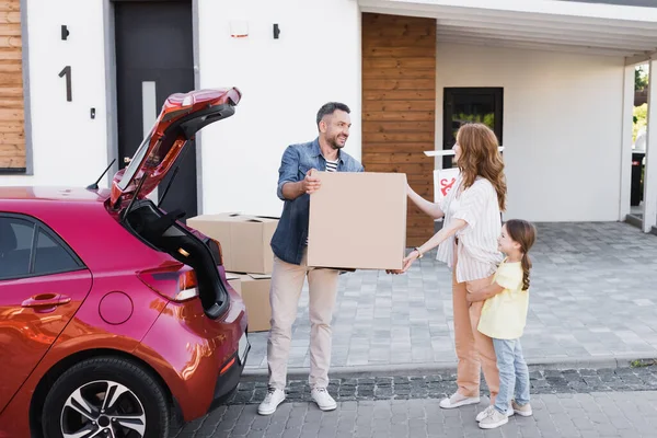 Full length of smiling wife taking carton box from husband while standing near daughter and car — Stock Photo
