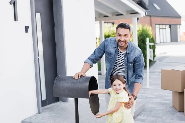 Fröhlicher Vater umarmt Tochter in der Nähe eines leeren Briefkastens, während er auf verschwommenem Hintergrund in die Kamera in der Nähe des Hauses blickt — Stockfoto