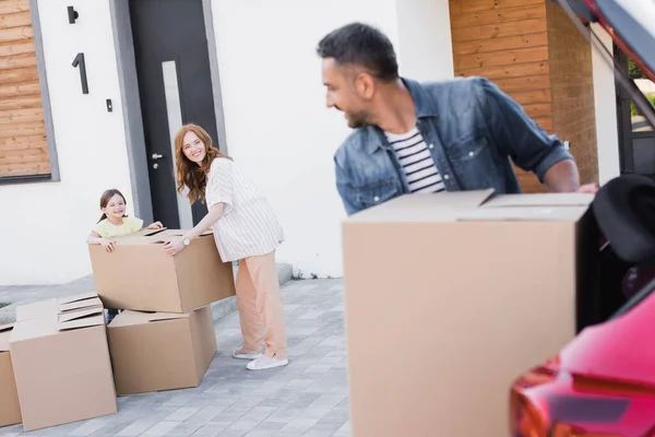 Happy mother and daughter near packages looking at blurred father with carton box near car trunk on foreground — Stock Photo