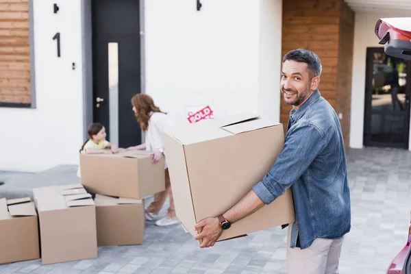 Glücklicher Ehemann mit Karton, der in die Kamera schaut, nahe verschwommener Frau und Tochter in der Nähe von Paketen im Hintergrund — Stockfoto