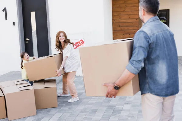 Happy woman and girl holding package while looking at blurred man with carton box on foreground — Stock Photo
