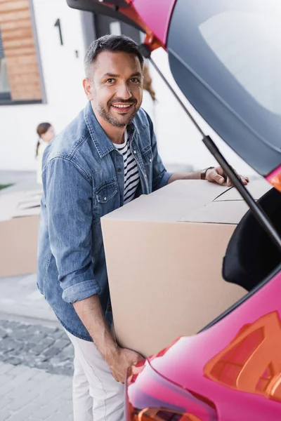 Hombre feliz mirando a la cámara mientras toma la caja de cartón del maletero del coche sobre un fondo borroso - foto de stock