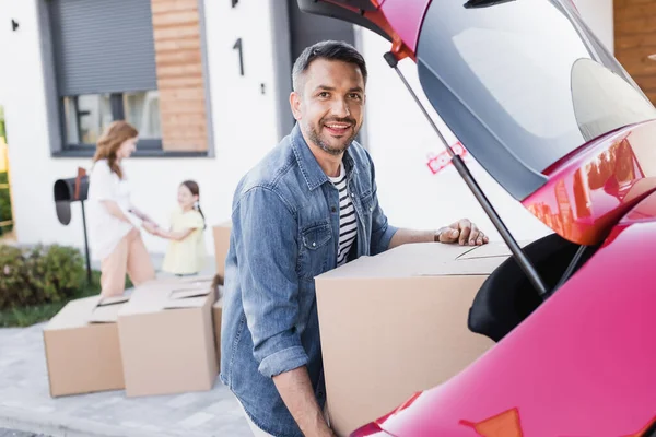 Hombre sonriente mirando a la cámara mientras toma la caja de cartón del maletero del coche con la mujer borrosa y la chica en el fondo - foto de stock