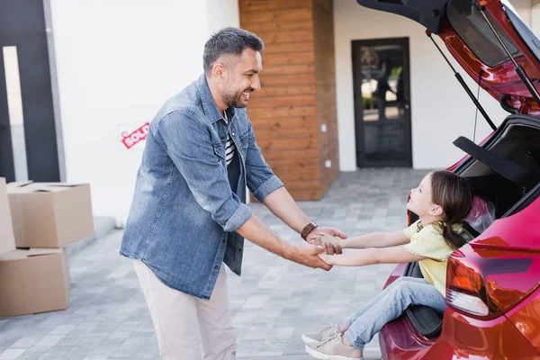 Sonriente hija sentada en el maletero del coche y cogida de la mano con el padre cerca de cajas de cartón borrosas y la casa en el fondo - foto de stock