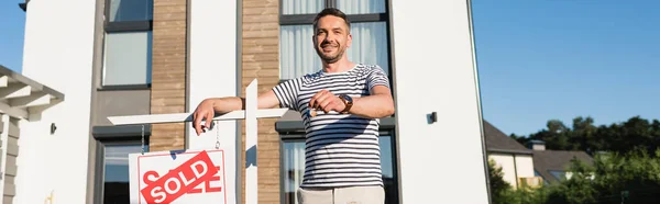 Uomo sorridente guardando la fotocamera mentre mostra la chiave vicino al segno con scritte vendute con casa moderna sullo sfondo, banner — Foto stock