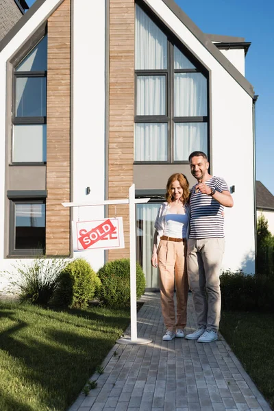 Full length of happy couple with key hugging and looking at camera while standing near modern house and sign with sold lettering — Stock Photo