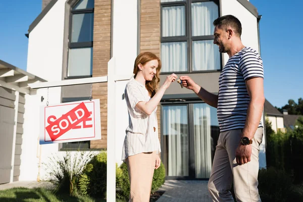 Femme heureuse prenant la clé de mari près signe avec lettrage vendu et maison moderne sur fond — Photo de stock