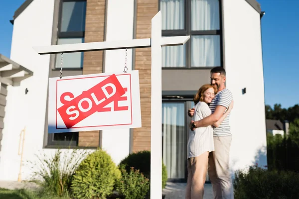 Schild mit verkauftem Schriftzug mit verschwommenem Paar, das sich in der Nähe des Hauses umarmt — Stockfoto