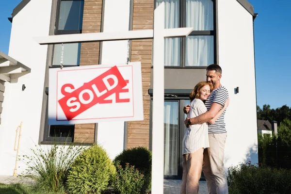 Happy couple embracing while standing near sign with sold lettering on foreground — Stock Photo