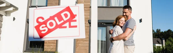 Smiling couple embracing while standing near sign with sold lettering on blurred foreground, banner — Stock Photo