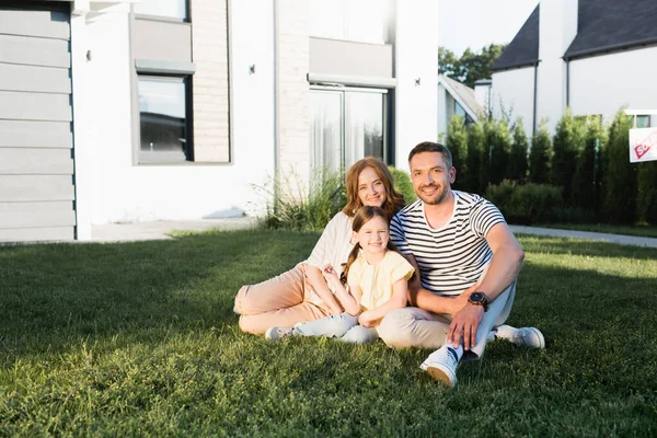 Glückliche Familie blickt in die Kamera, während sie auf dem Rasen neben modernem Haus sitzt — Stockfoto